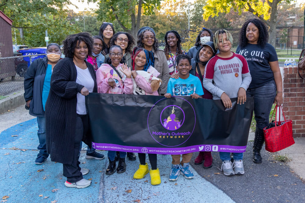 A group of Black women behind a Mother's Outreach Network banner smiling.