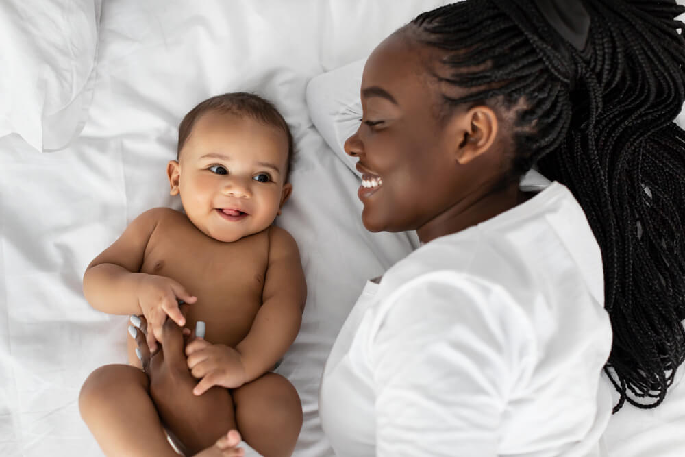 Young Black mother lying in bed with her infant child and smiling.